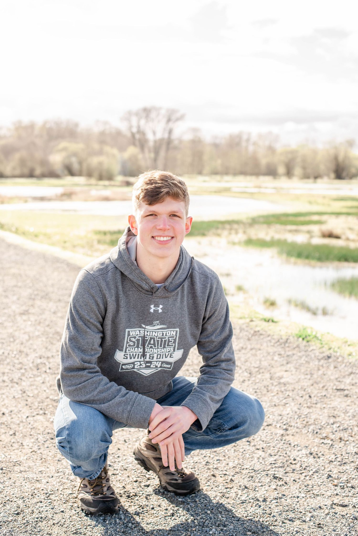 Person wearing grey hoodie squatting on gravel pathway with natural landscape behind.