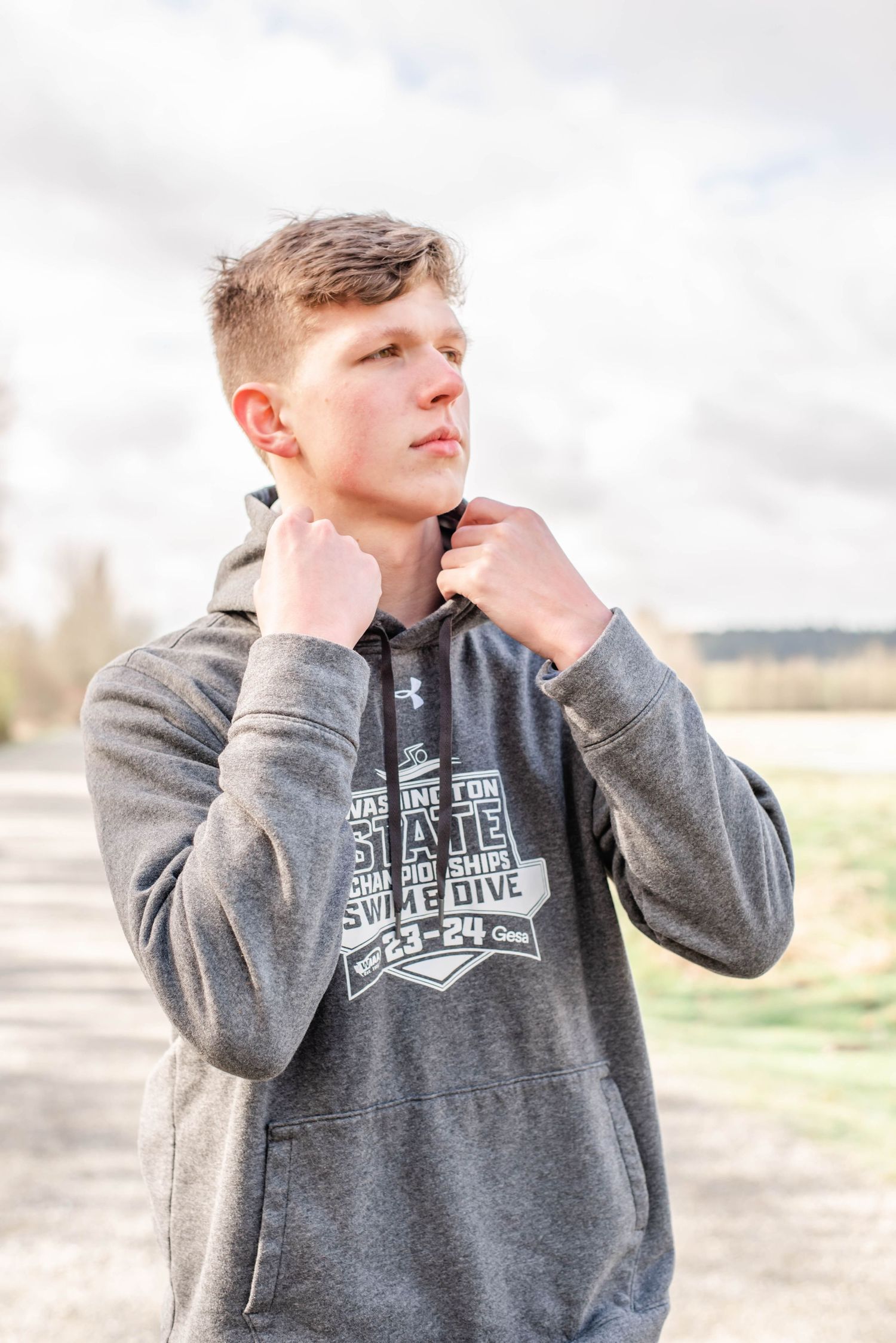 Person in grey branded hoodie adjusting collar against cloudy sky.
