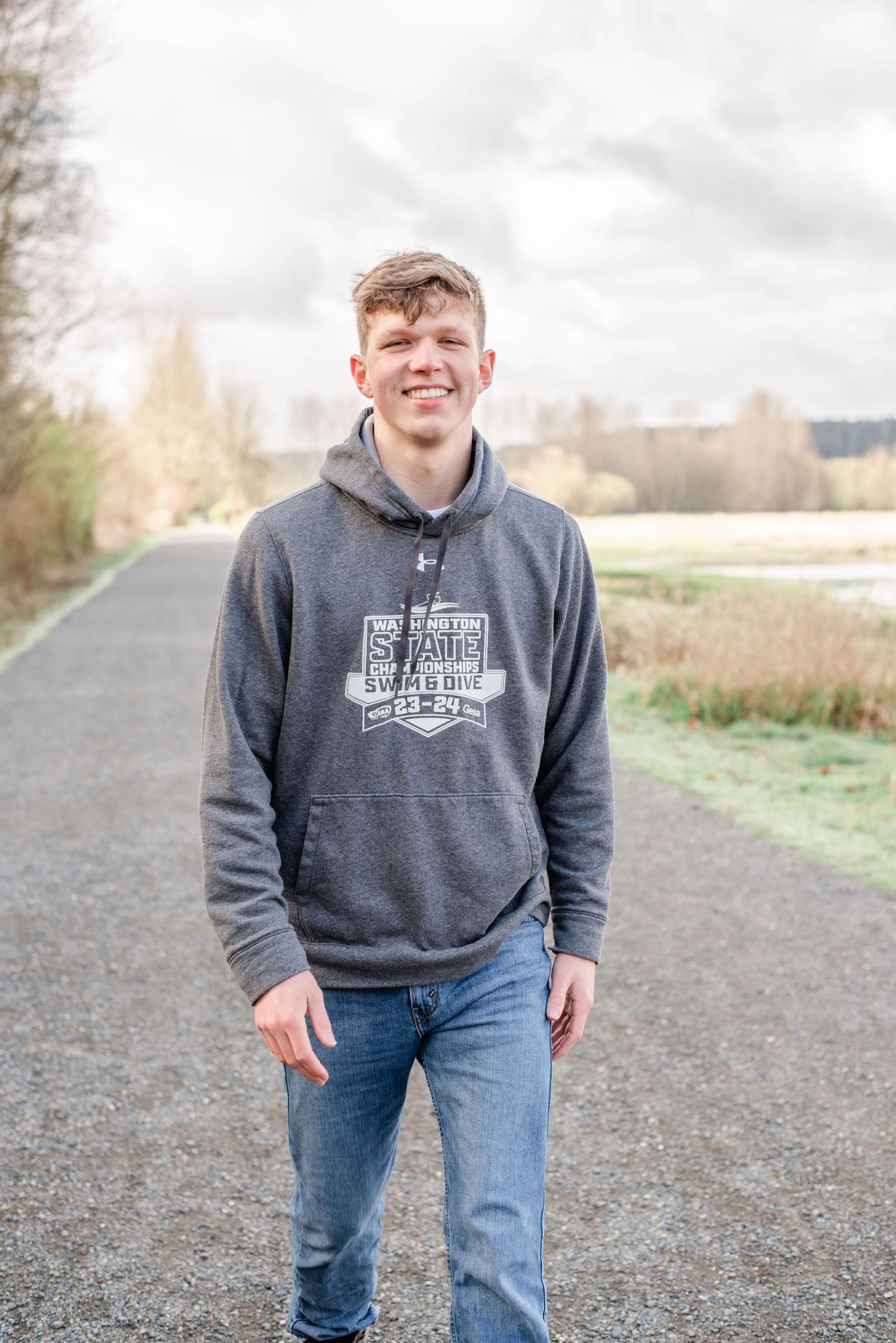 Person wearing grey hoodie standing on path with rural landscape background.