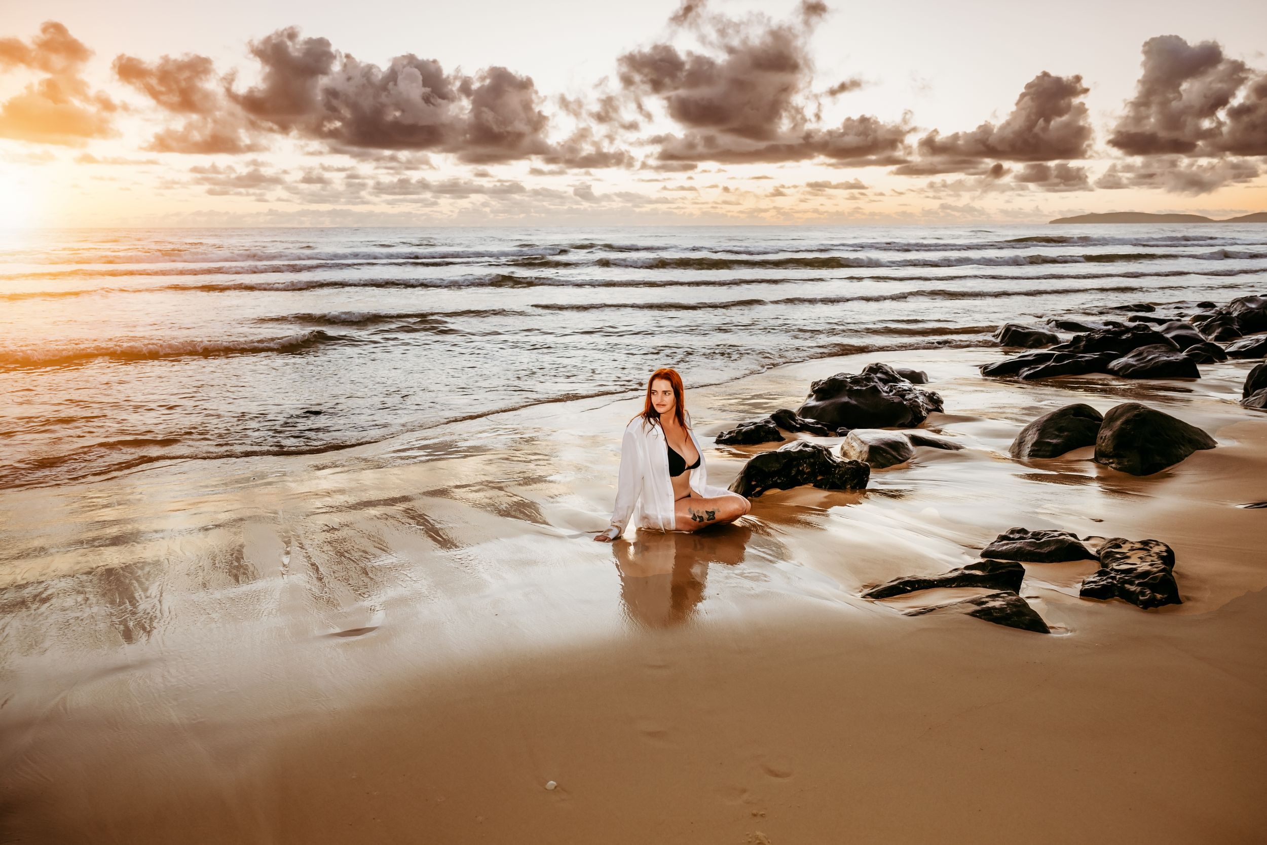Person in white dress sitting on wet beach sand among rocks at sunset.