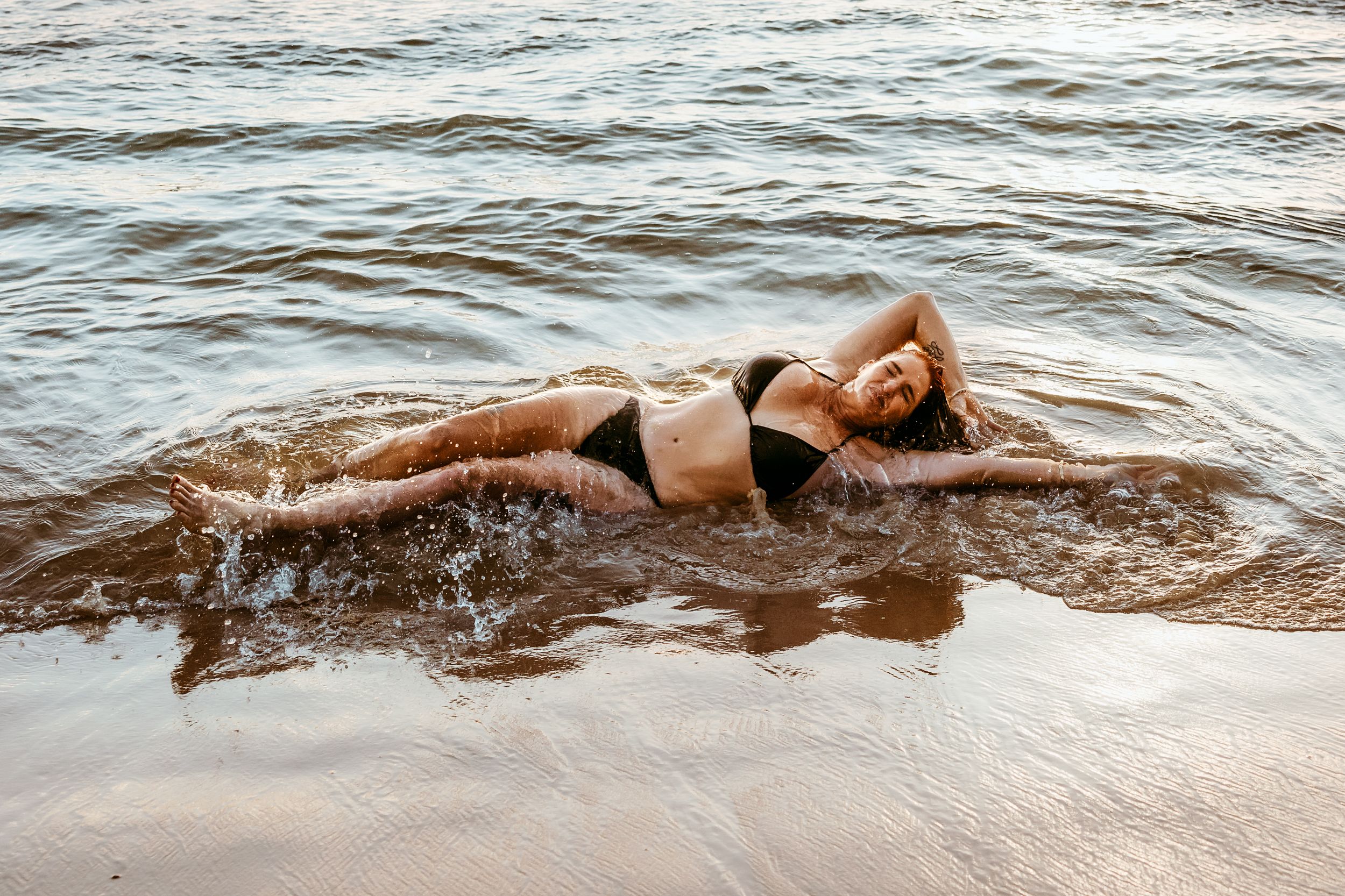 Person in black swimwear lying in shallow ocean water on sandy beach at sunset.