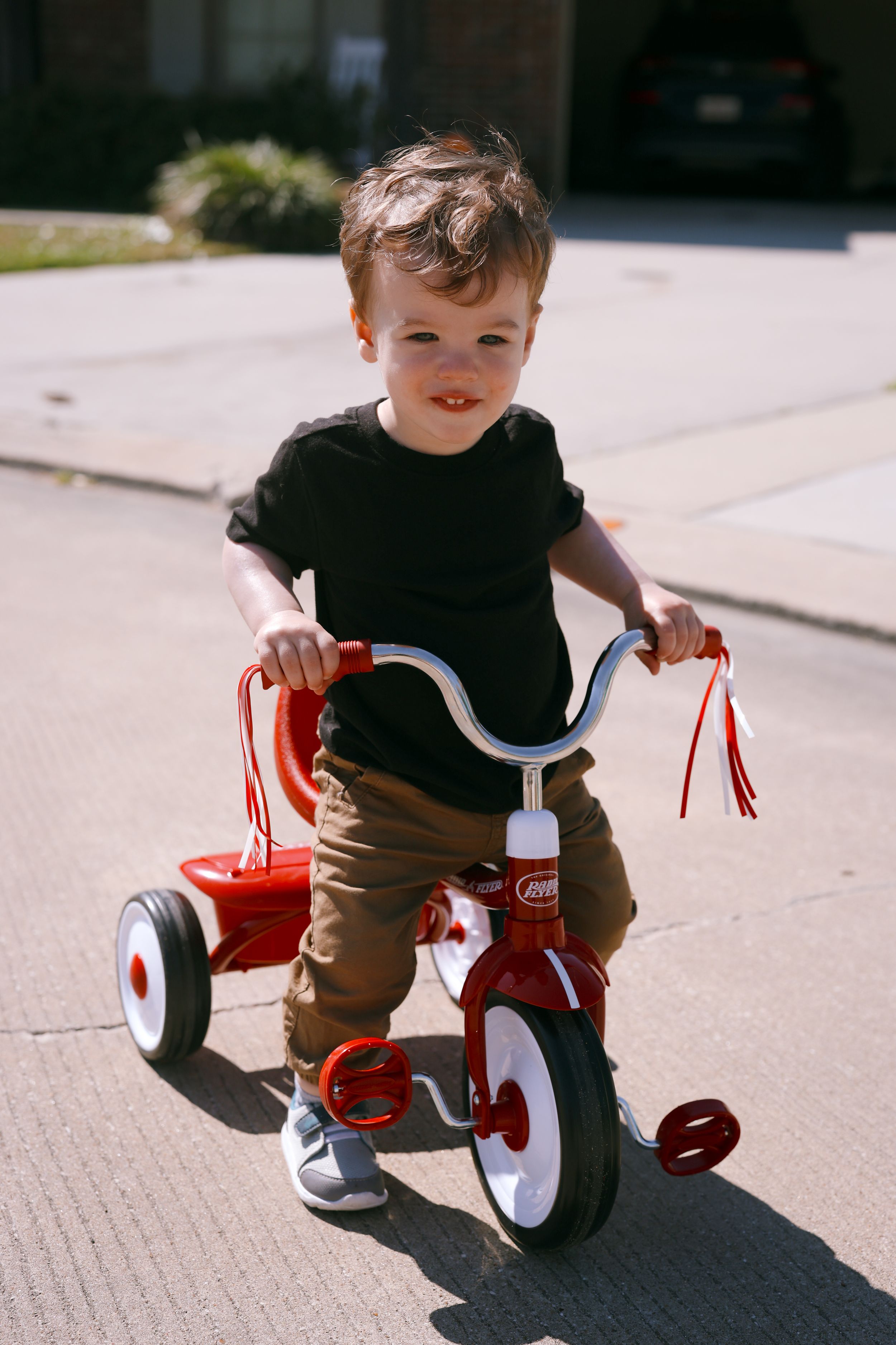 A small child rides a classic red tricycle on a sunny sidewalk while wearing a black shirt and brown pants.