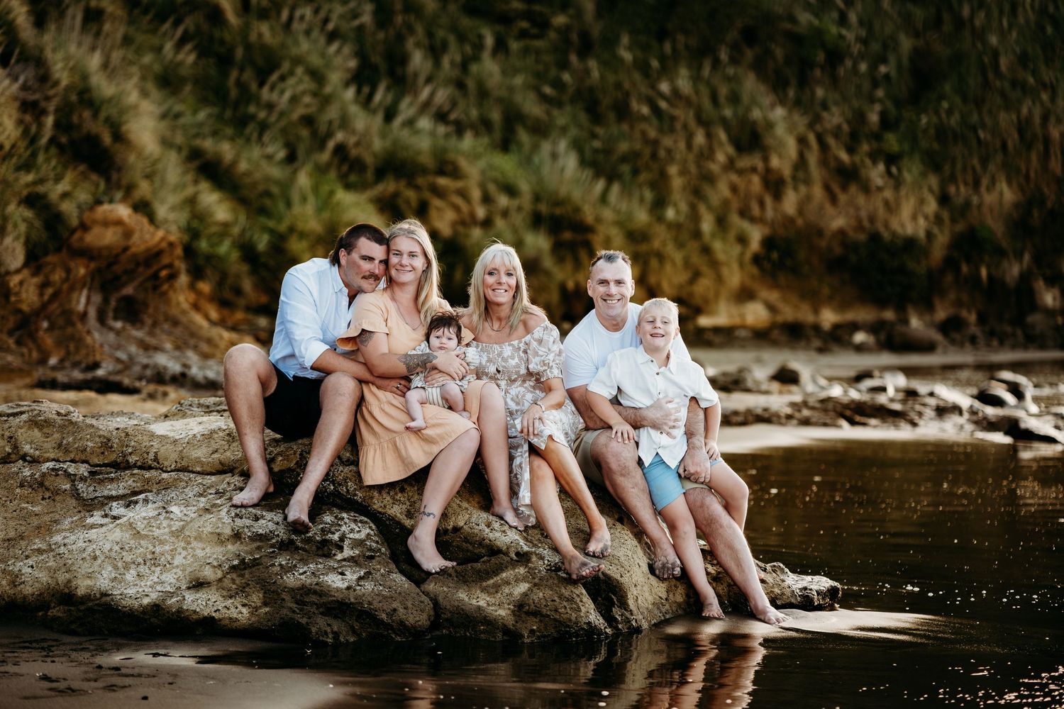 Family sitting together on rocky shore with feet dangling in water.