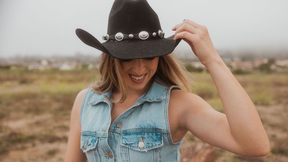 A person wearing a denim vest and cowboy hat with pearl details poses against an arid landscape.
