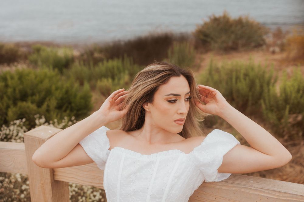 A person in a white off-shoulder dress poses by a wooden fence near coastal cliffs.