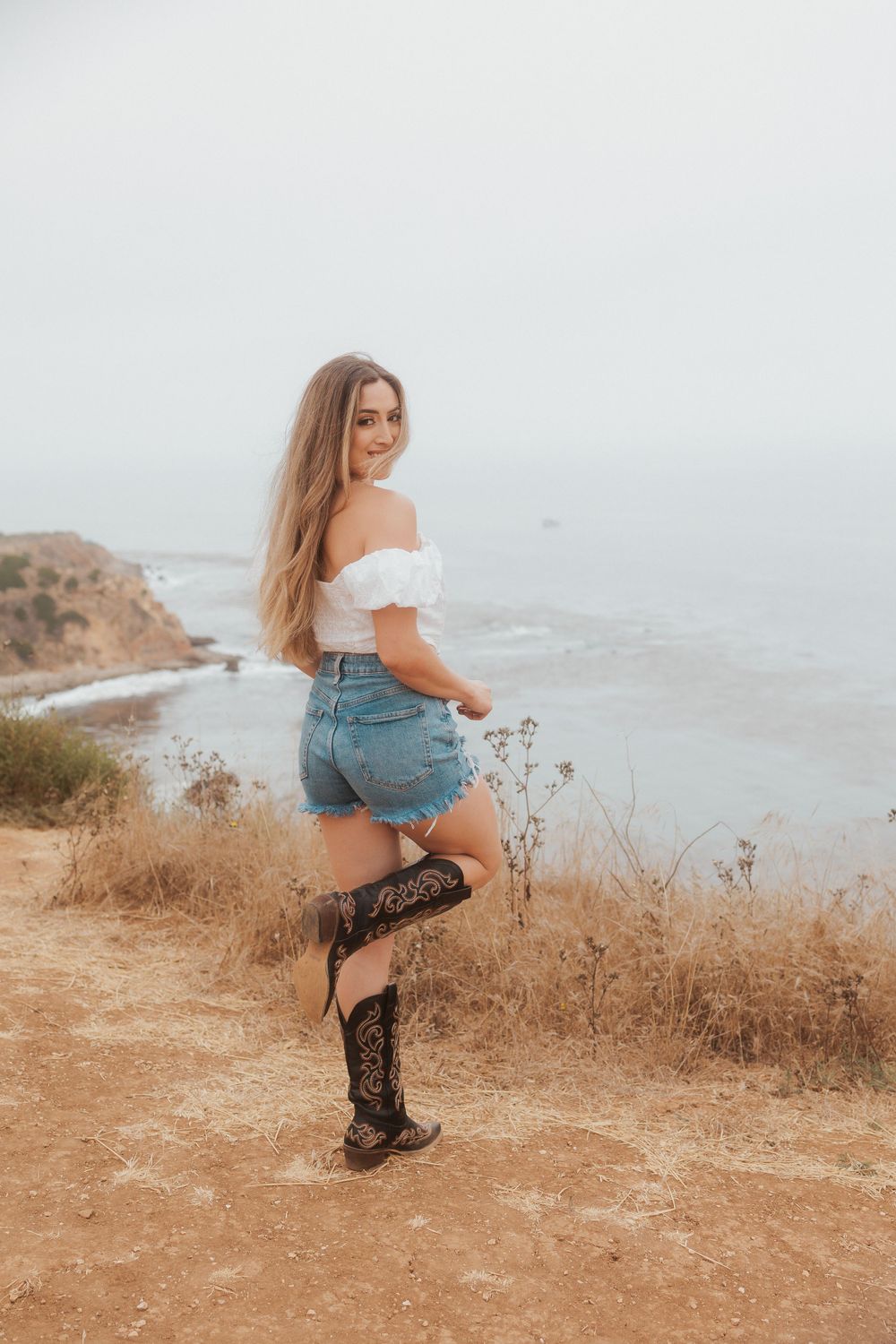 A person in western boots and denim shorts poses on a coastal trail with ocean views.