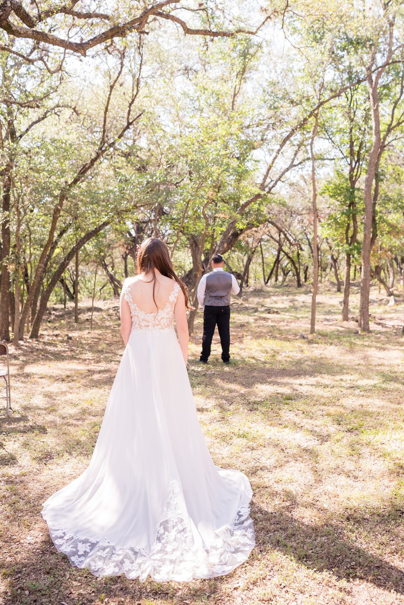 Bride in lace-detailed gown awaits first look in scenic wooded setting.