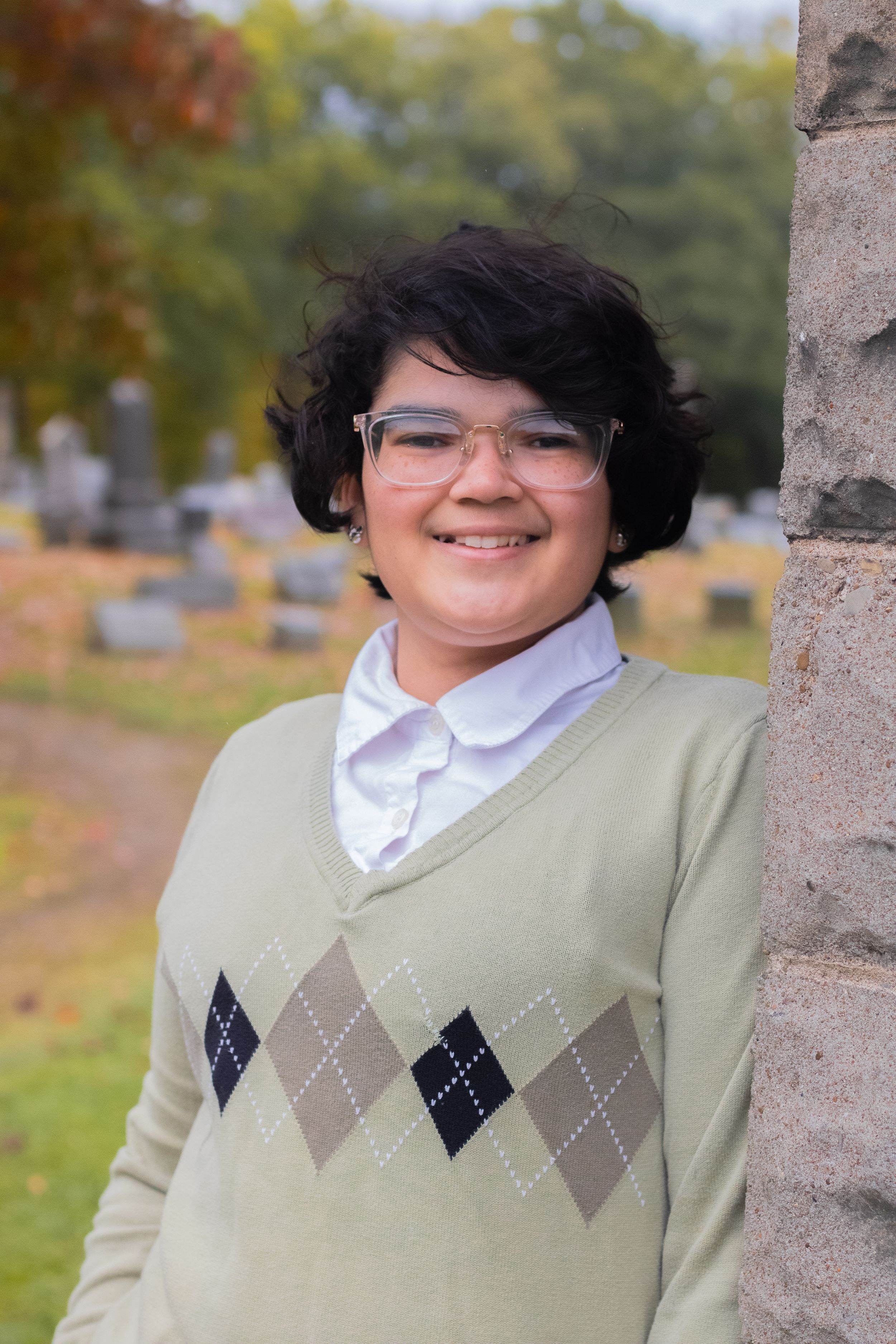 A cheerful portrait of someone in an argyle sweater vest and glasses against autumn foliage.