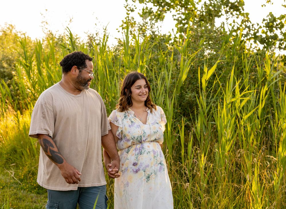 A couple walks through tall grass in a sunlit field during golden hour.
