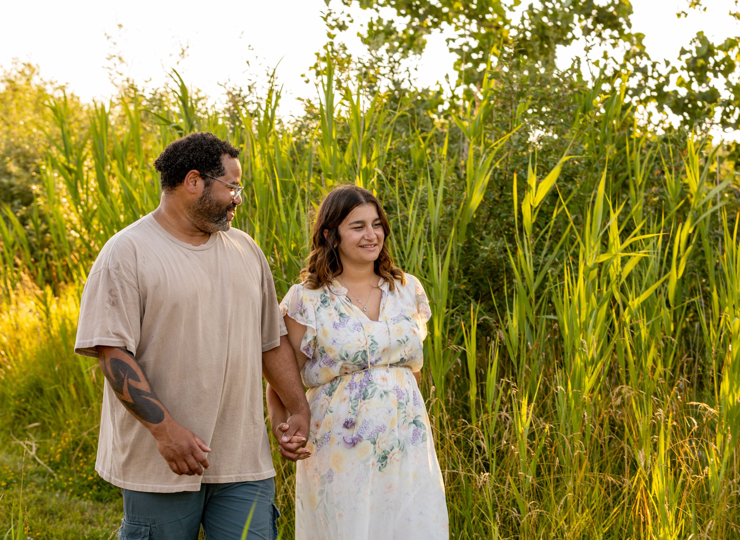 A couple walks through tall grass in a sunlit field during golden hour.