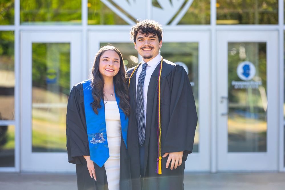 Two smiling graduates in black caps and gowns pose outside a university building on graduation day.
