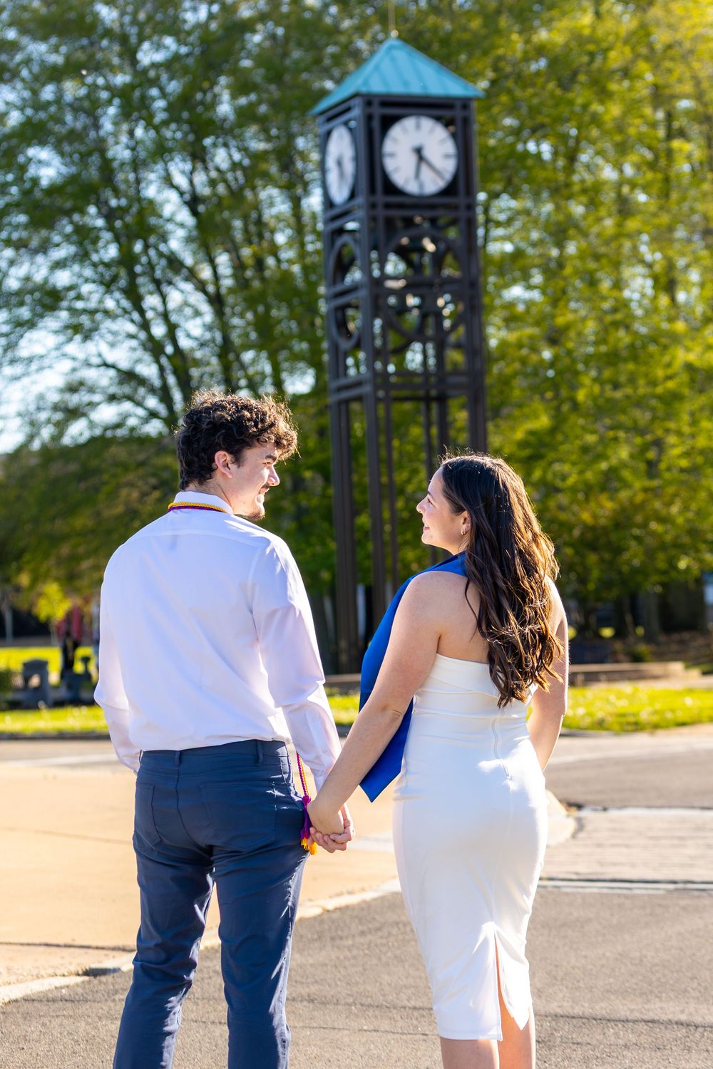 Couple holding hands walking toward a black clock tower surrounded by green trees on a sunny day.