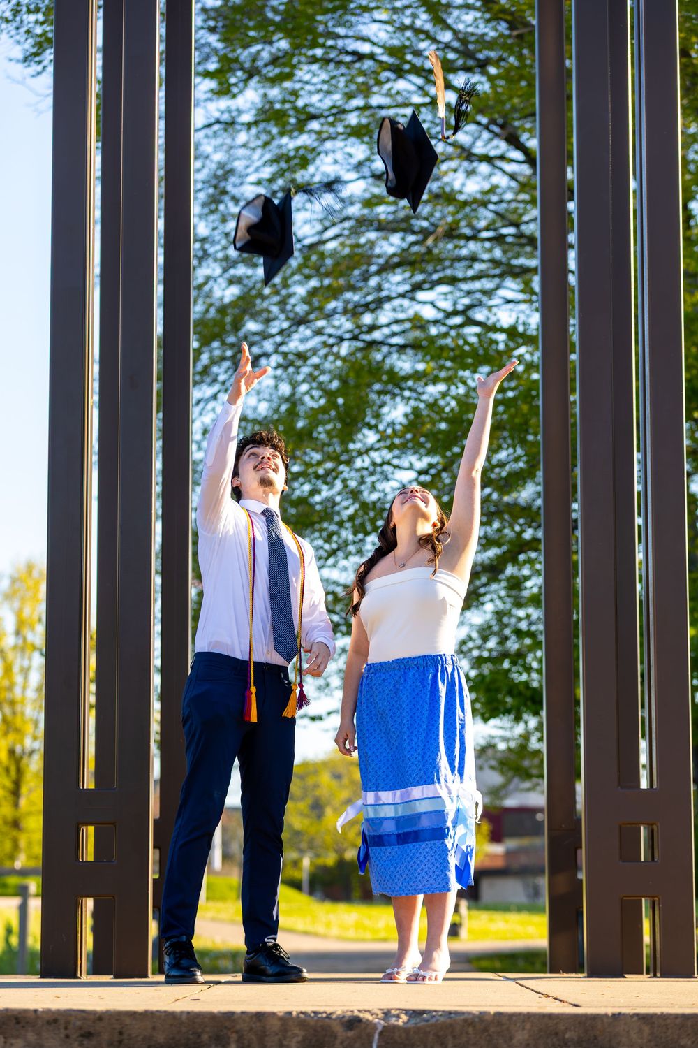 Two graduates in formal attire joyfully toss their black graduation caps into the air outdoors on a sunny day.