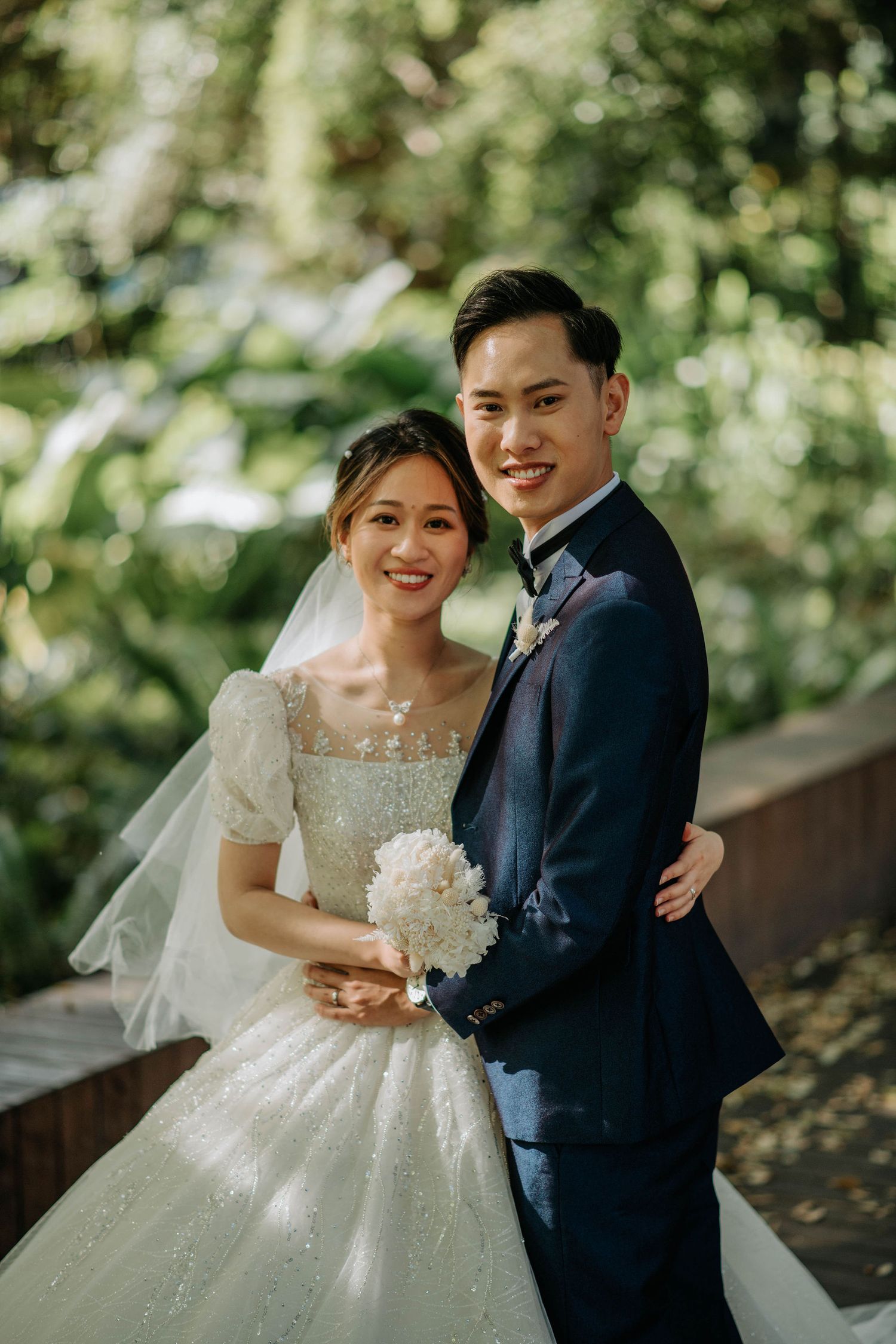 Elegant couple in wedding attire pose together in a sunlit garden setting.