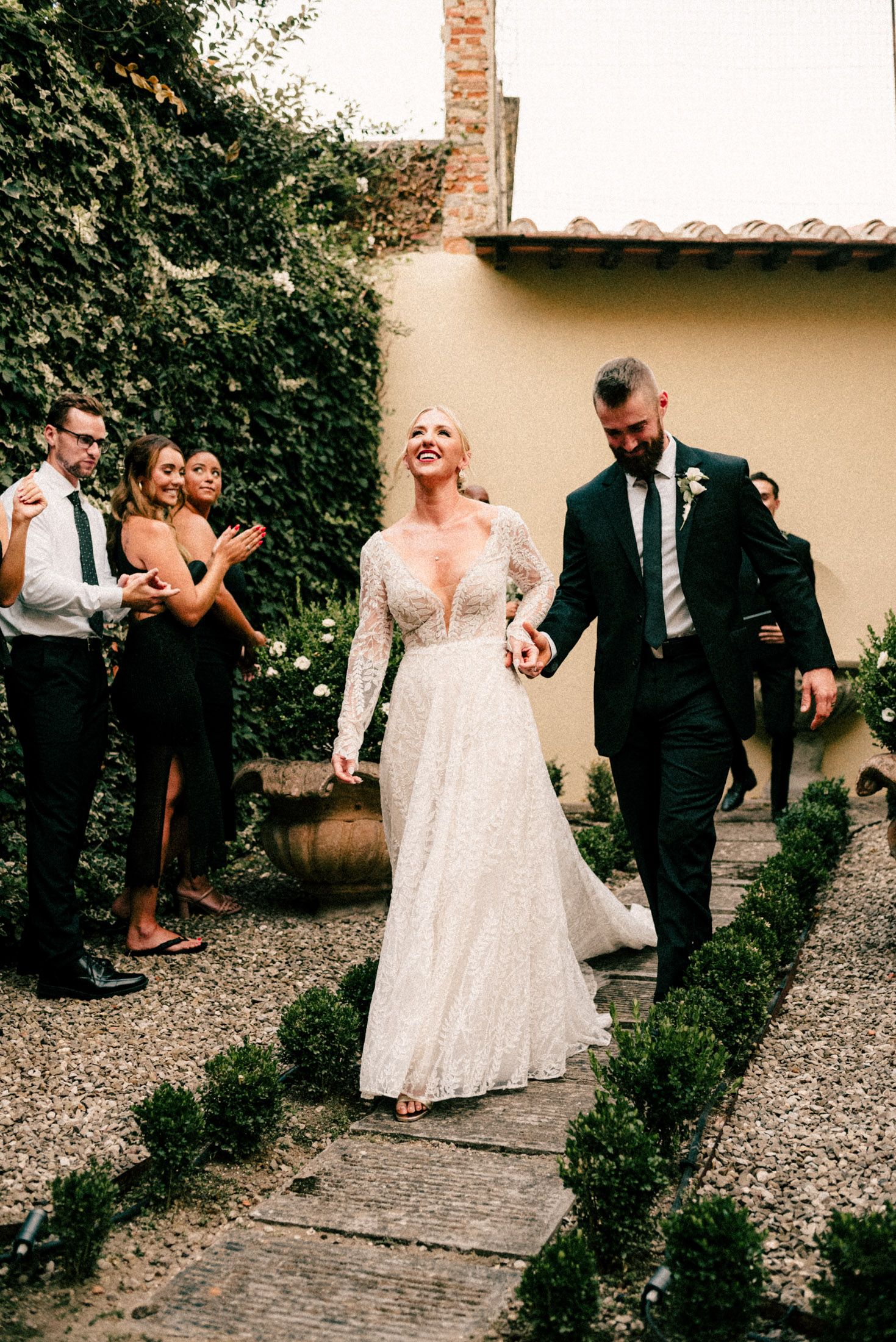 A wedding couple walks down a garden path while guests clap and celebrate their union.