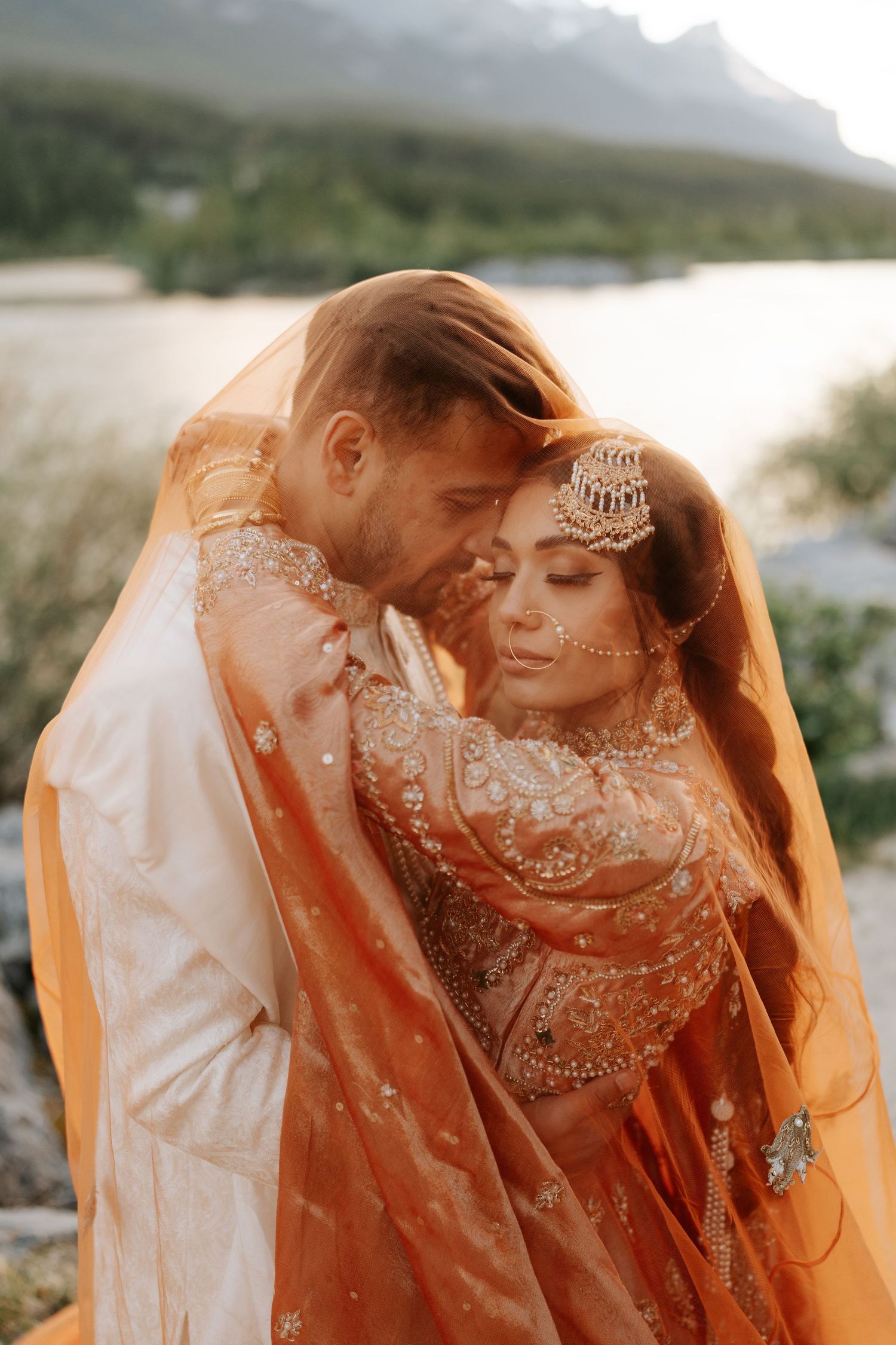 Couple embracing under an orange veil in front of scenic mountain landscape.