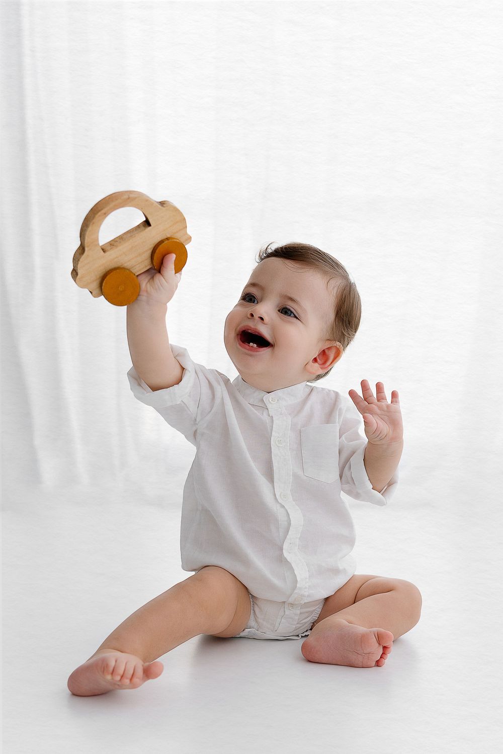 Baby in white outfit plays with wooden toy car on white background.