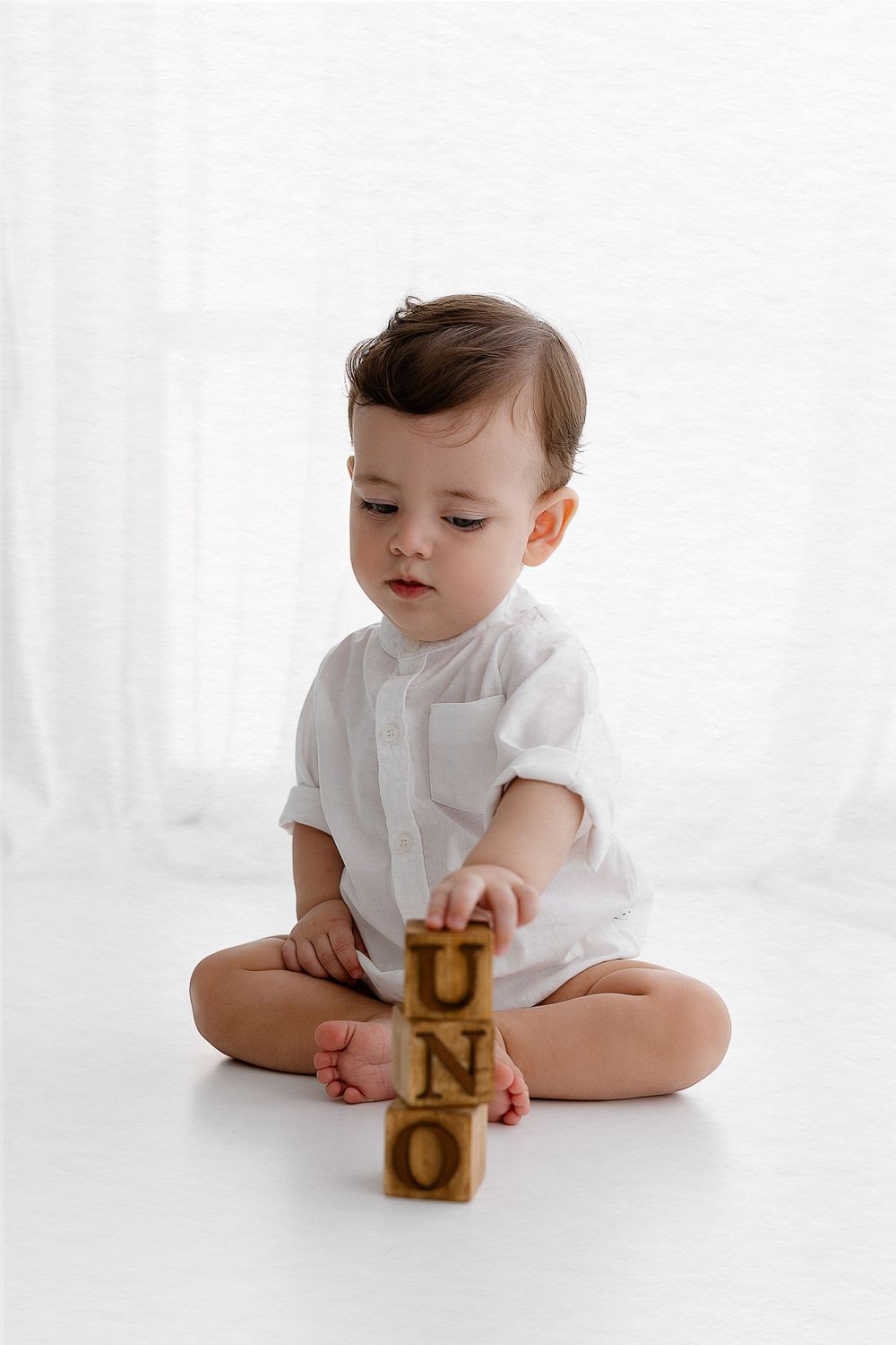 Baby in white shirt stacks wooden blocks on white floor.