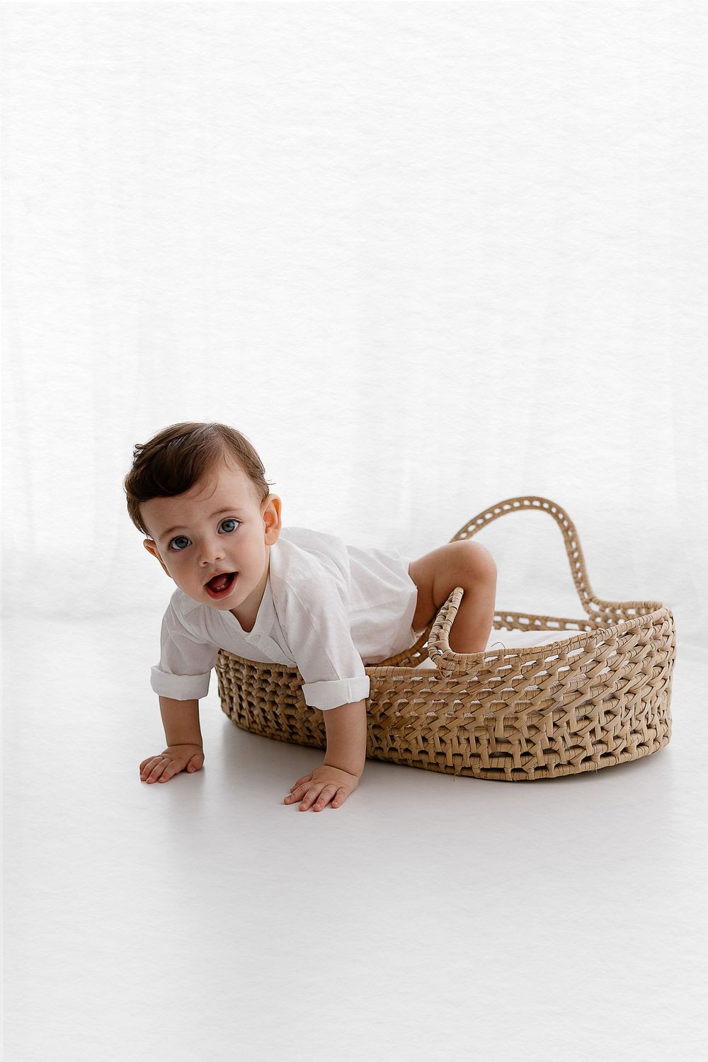 Baby in white crawls out of woven basket on white background.