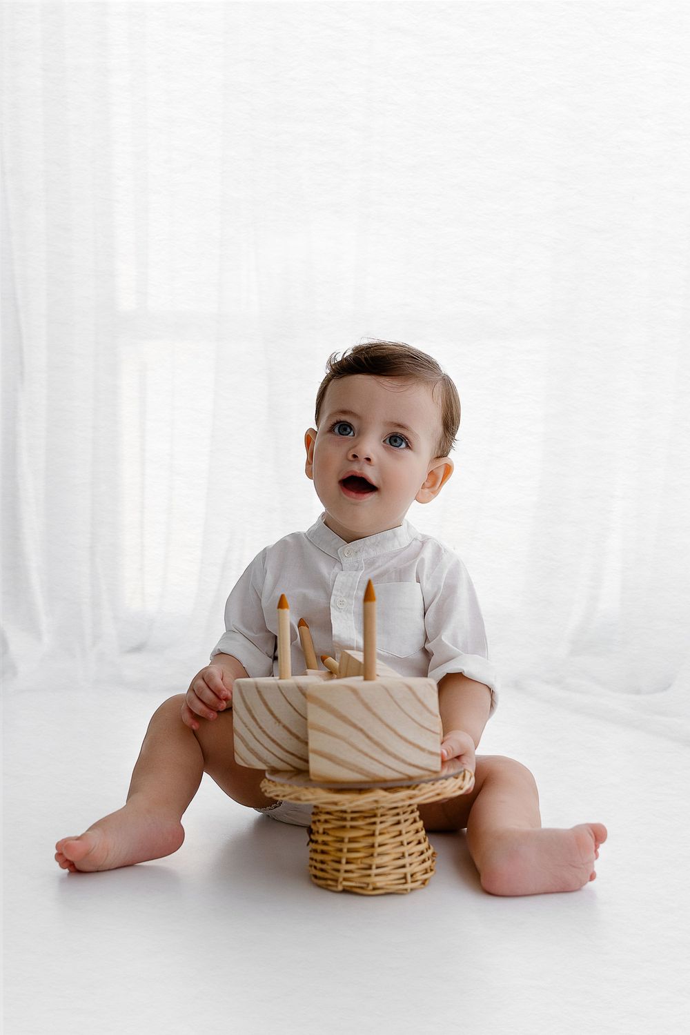 A baby in white sits with a wooden birthday cake decoration on a white background.