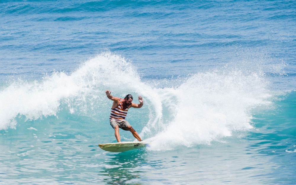 Surfer rides along a breaking wave in a striped shirt against bright blue ocean waters.