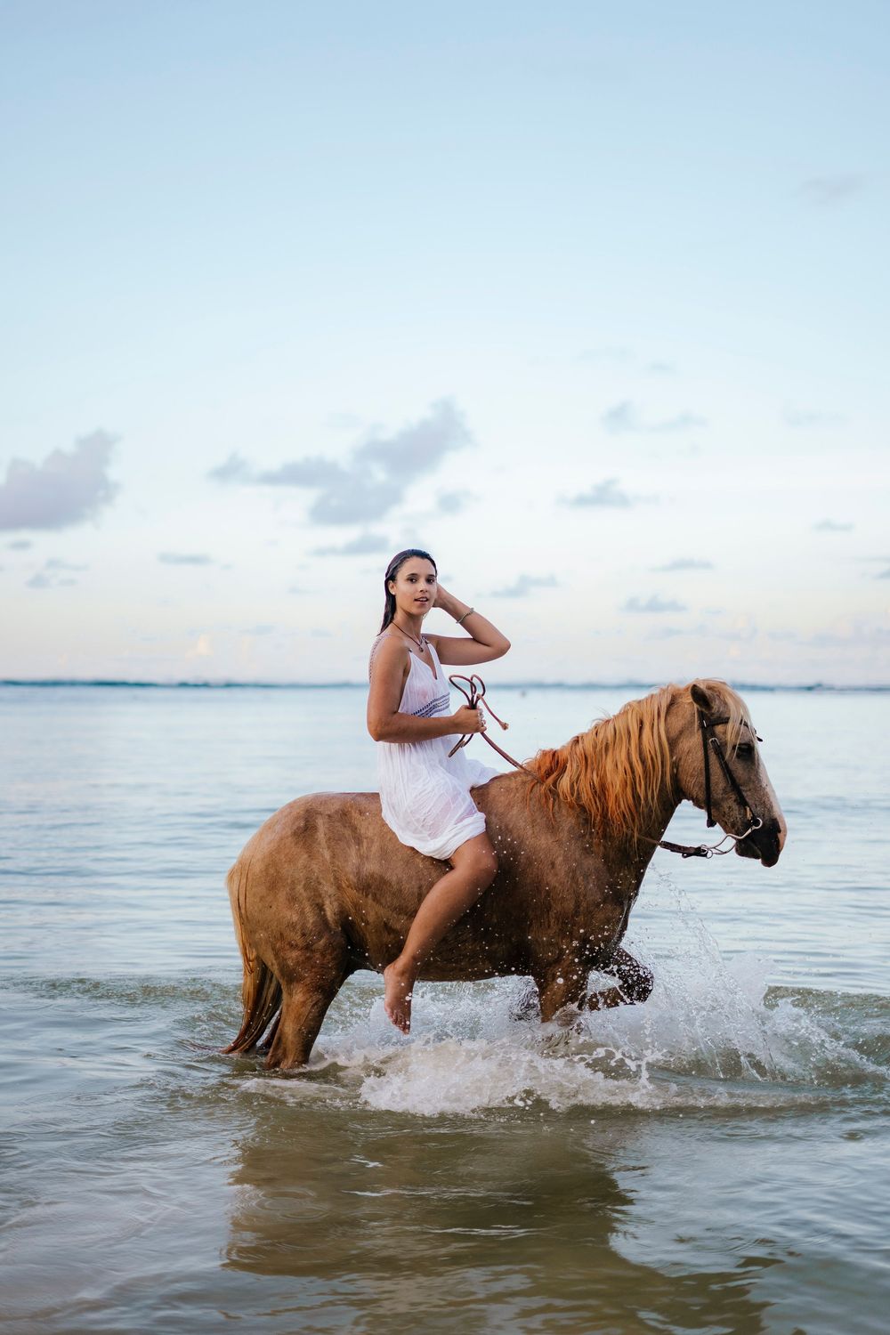 A horse and rider splash through shallow ocean waters at sunset creating a dynamic scene of motion and water spray.
