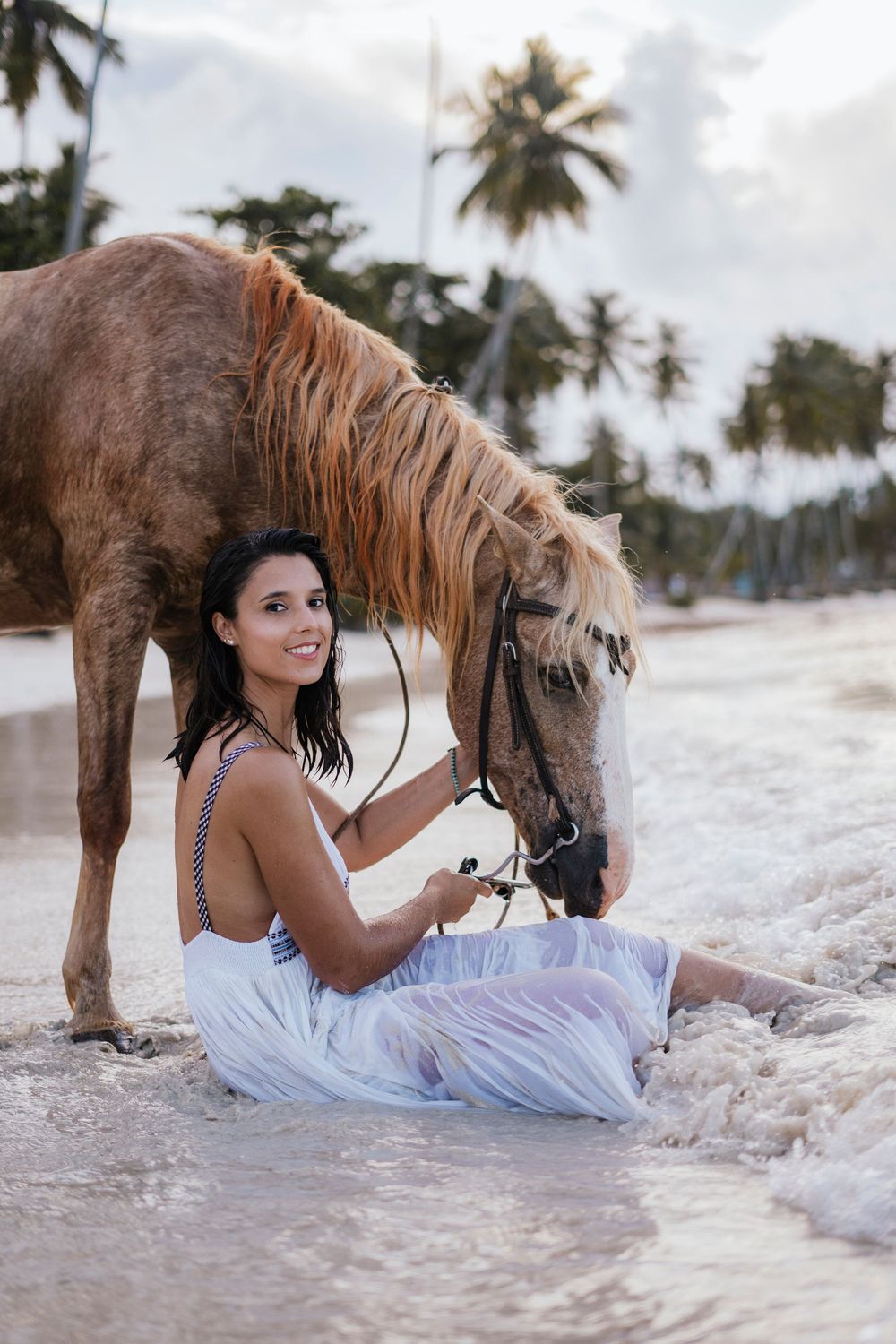 A brown horse and person sit together on a tropical beach with palm trees in the background.