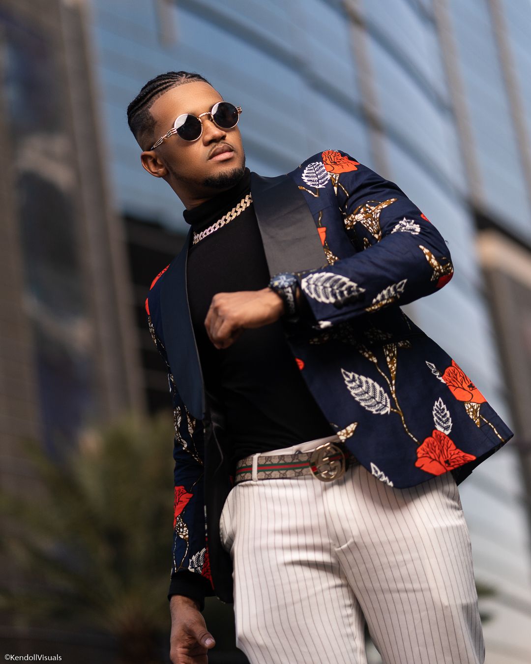 Stylish man posing in a floral navy blazer, white pinstripe pants, and round sunglasses outdoors.