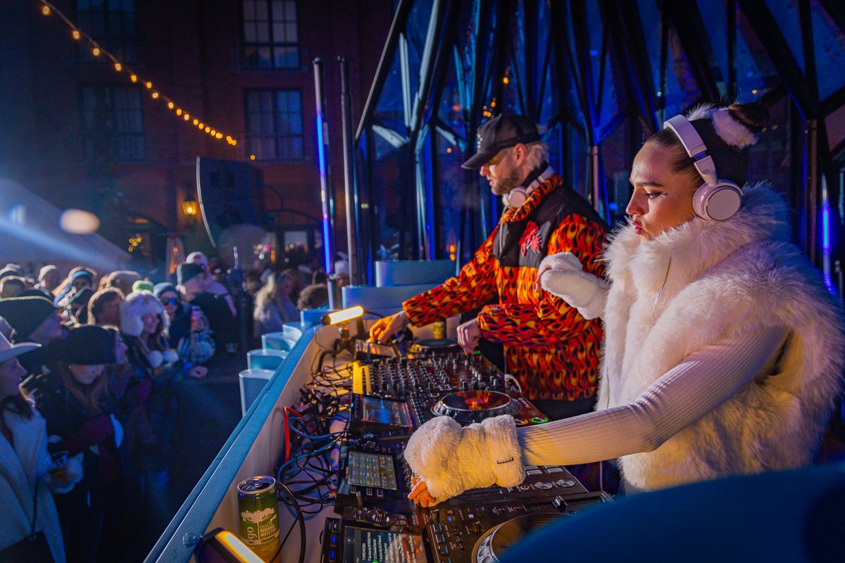 DJs performing at nighttime outdoor event with string lights and blue backdrop.