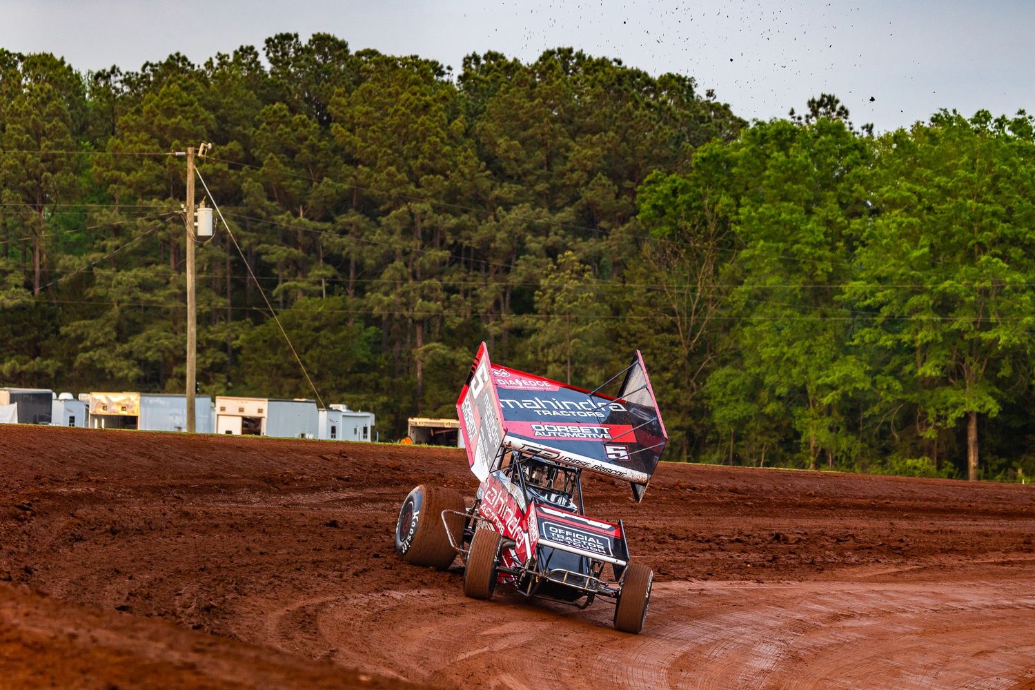 Dirt track racing scene with multiple sprint cars competing on a red clay oval under evening light.
