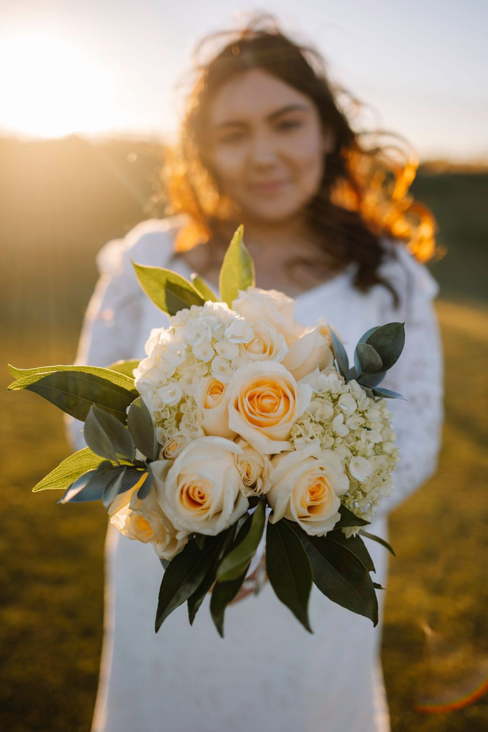 Wedding bouquet of peach roses and white hydrangeas held in sunlit field during golden hour.