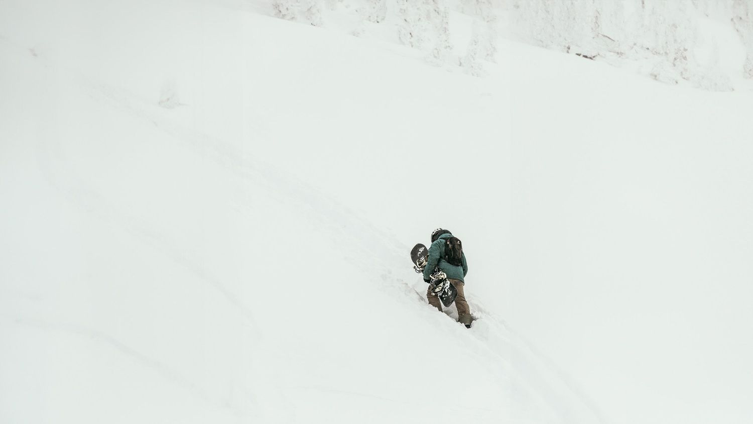 A snowboarder hiking up a mountain in knee deep snow while carrying his gear at the Revelstoke Ski Resort in Banff Canada.