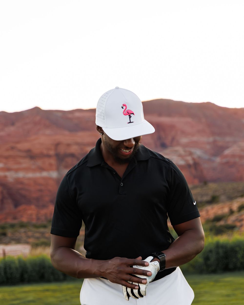 Man in black polo and white flamingo cap smiling on a golf course with red rock formations in background.
