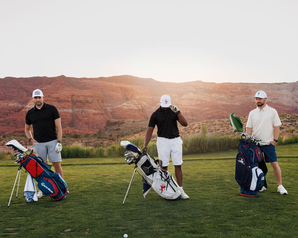 Three golfers with bags on a lush green course surrounded by stunning red rock canyon landscape at sunset.