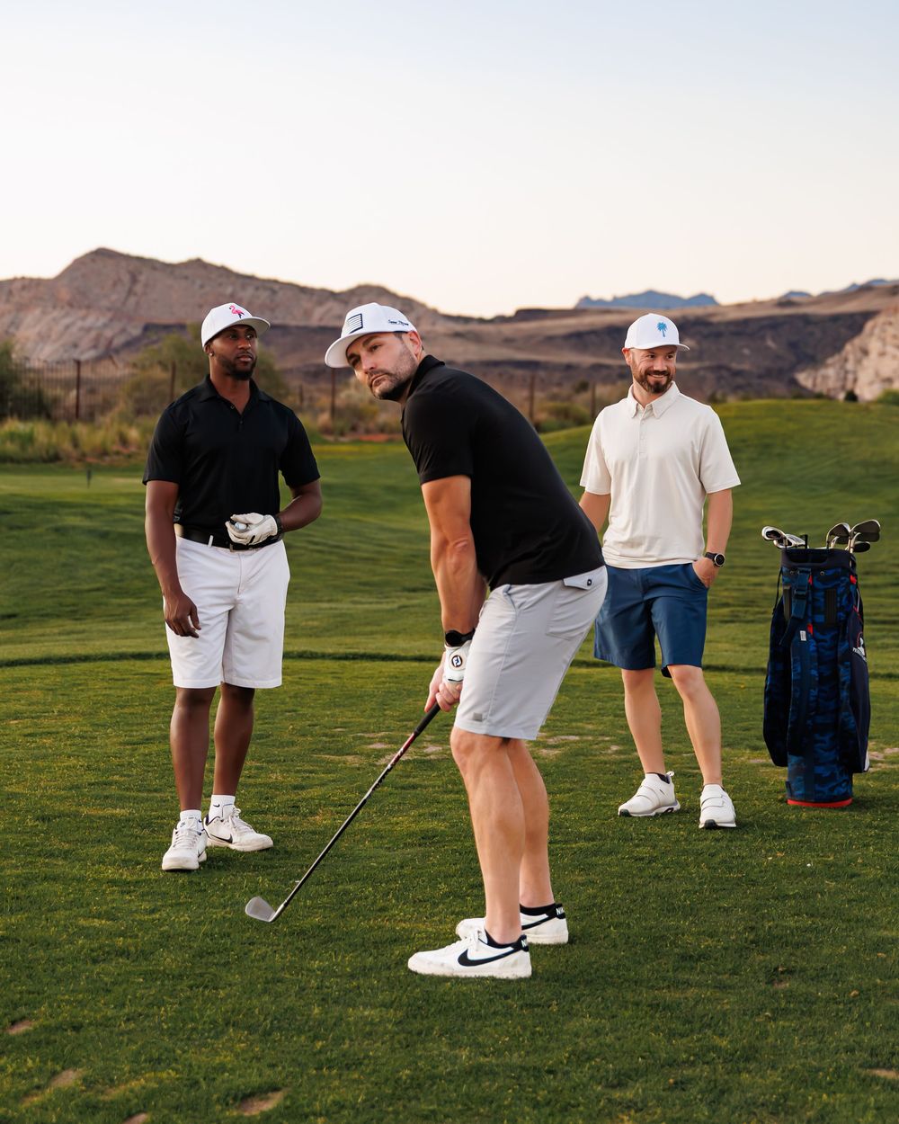 Three men playing golf on a scenic desert course, one mid-swing with Nike shoes, others watching nearby.