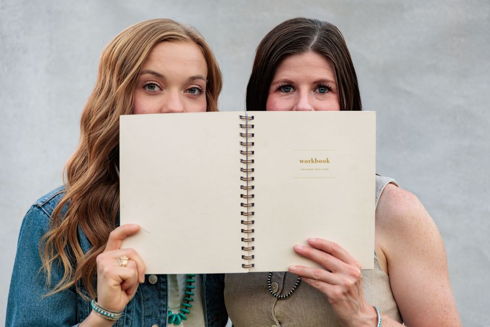 Two women holding open a spiral-bound notebook in front of their faces against a gray background.