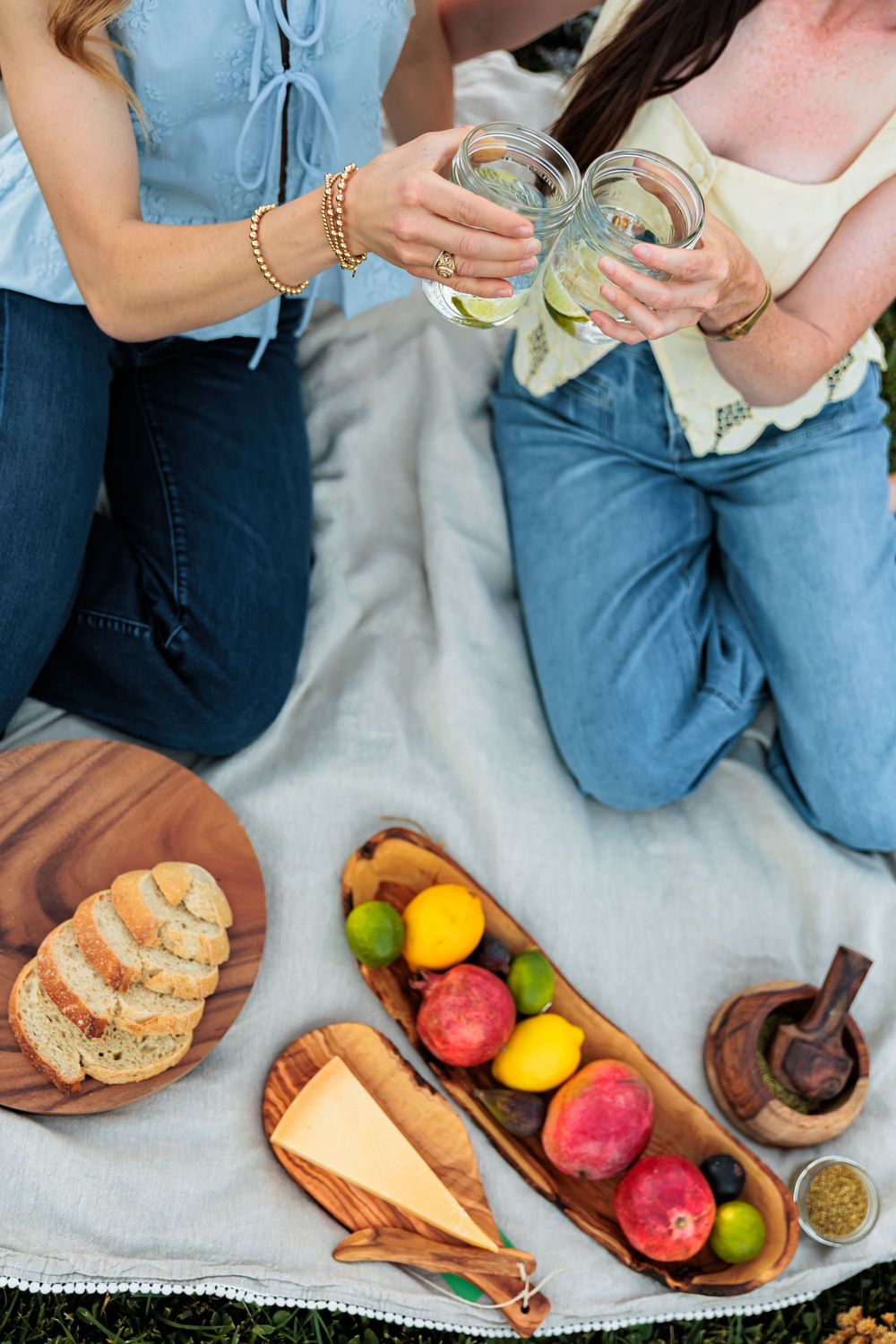 Two women toasting mason jar drinks at a picnic with fruit, bread, and cheese on a blanket outdoors.