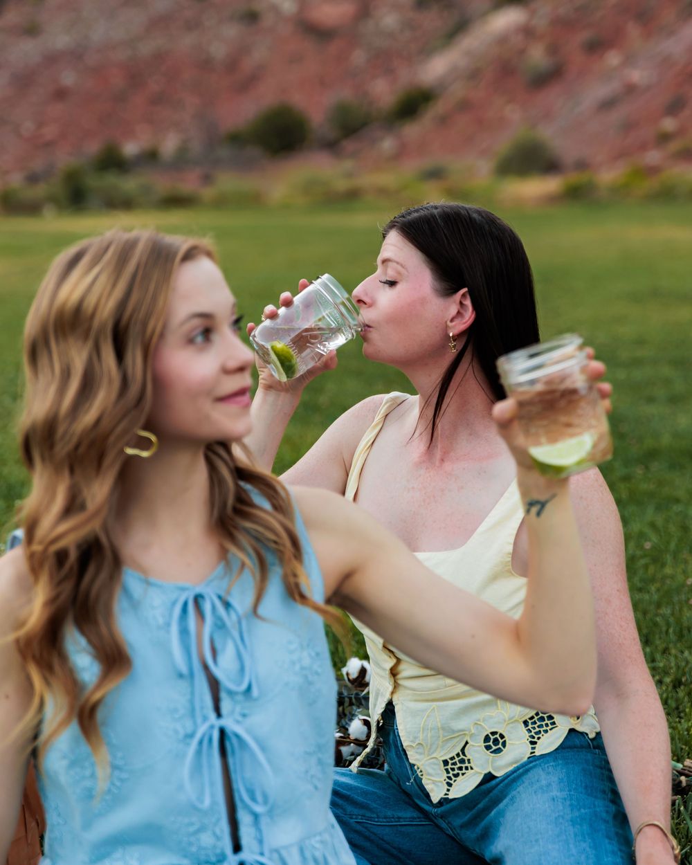 Two women sitting outdoors drinking from mason jars with green grass and red rock mountains behind them.