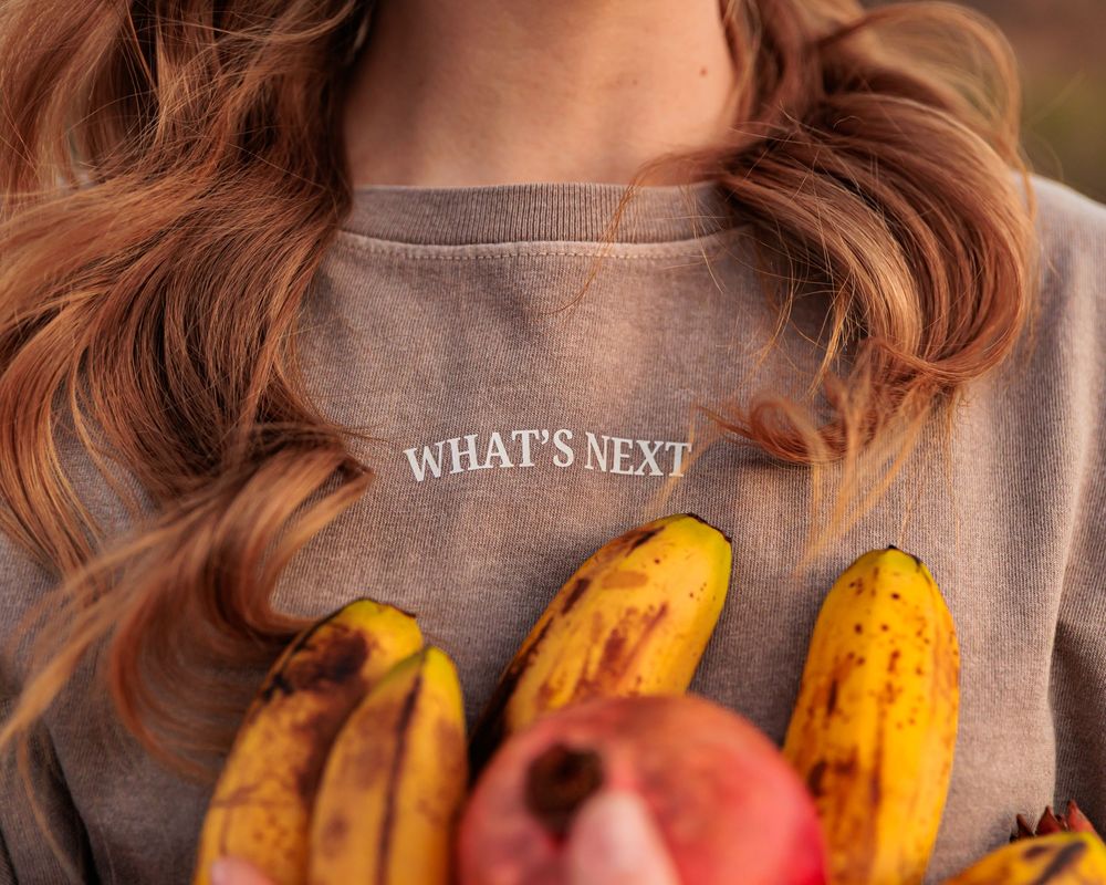 Woman in gray 'What's Next' sweatshirt holding ripe bananas and a pomegranate.