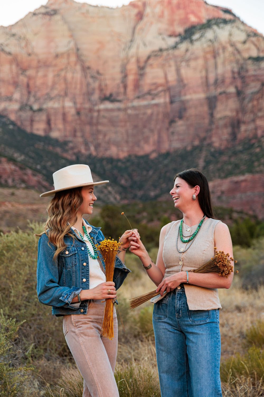 Two women smiling and holding wildflowers with red rock canyon mountains in the background.