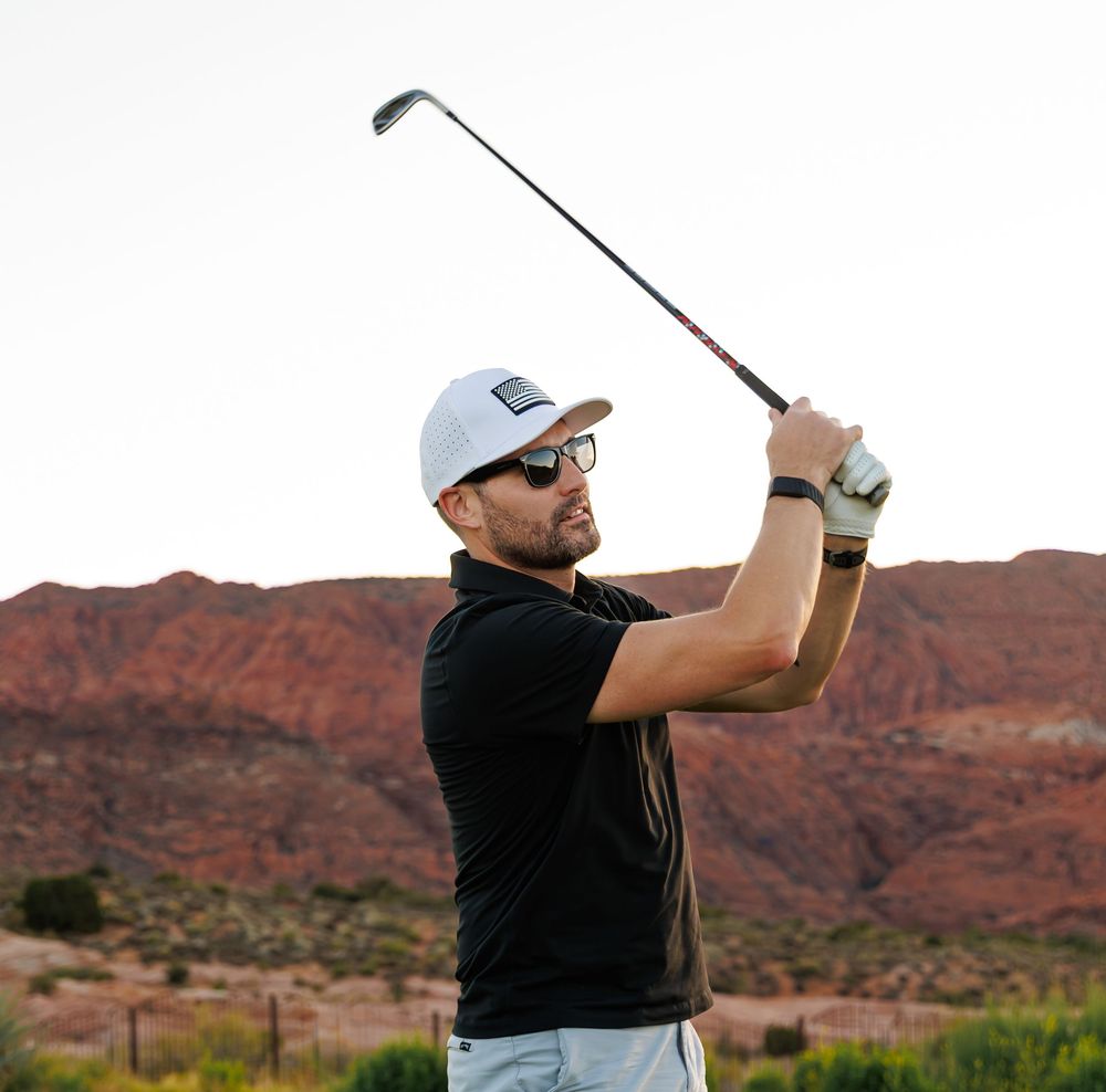 Man in black polo and white cap swings golf club on scenic red rock desert course.