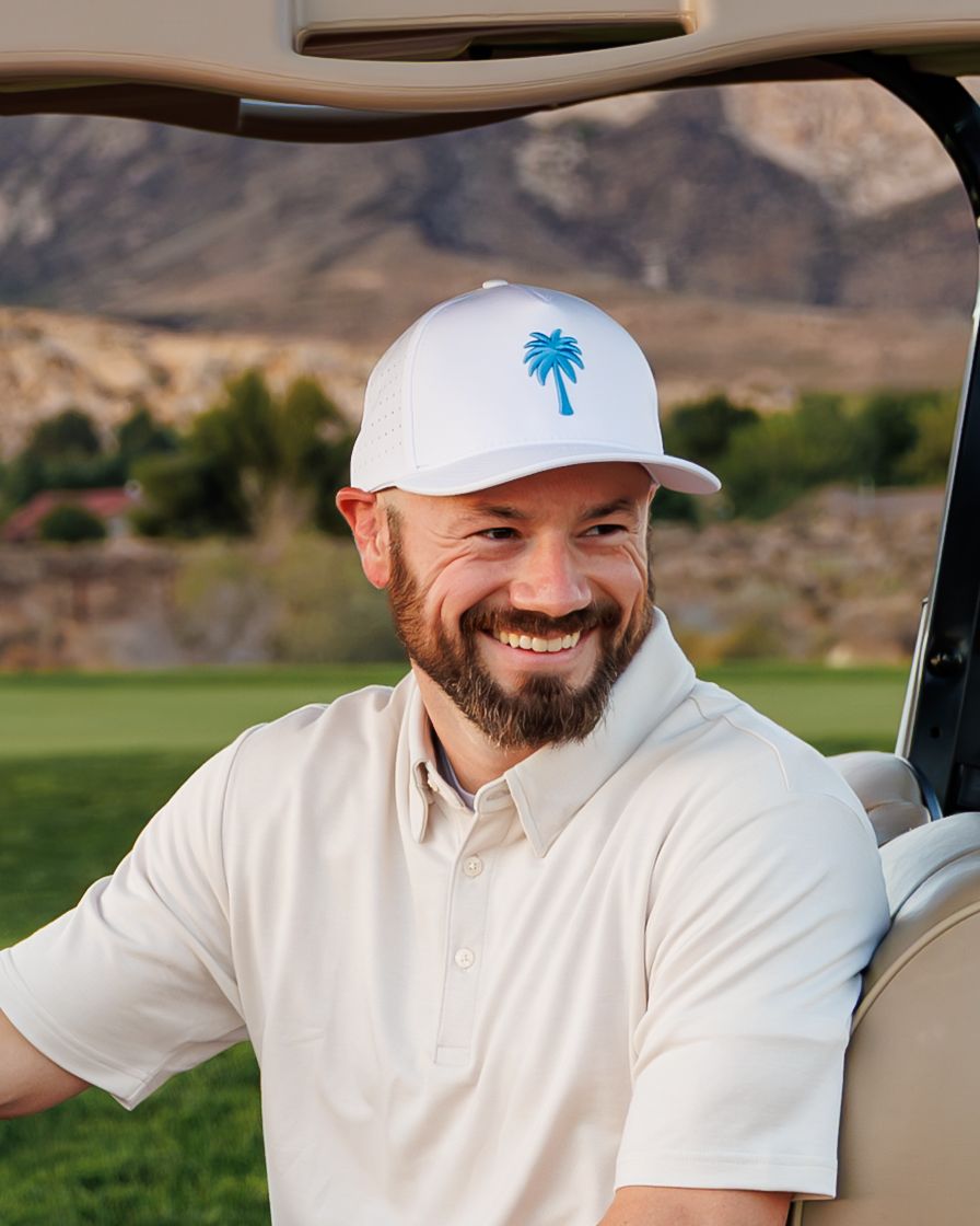 Smiling man with beard wearing white polo and palm tree cap sitting in golf cart on scenic mountain course.