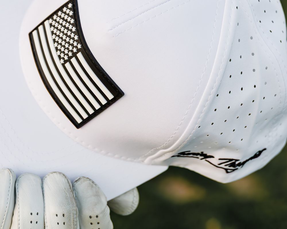 White golf hat with black American flag patch, held by a golfer wearing a white golf glove.