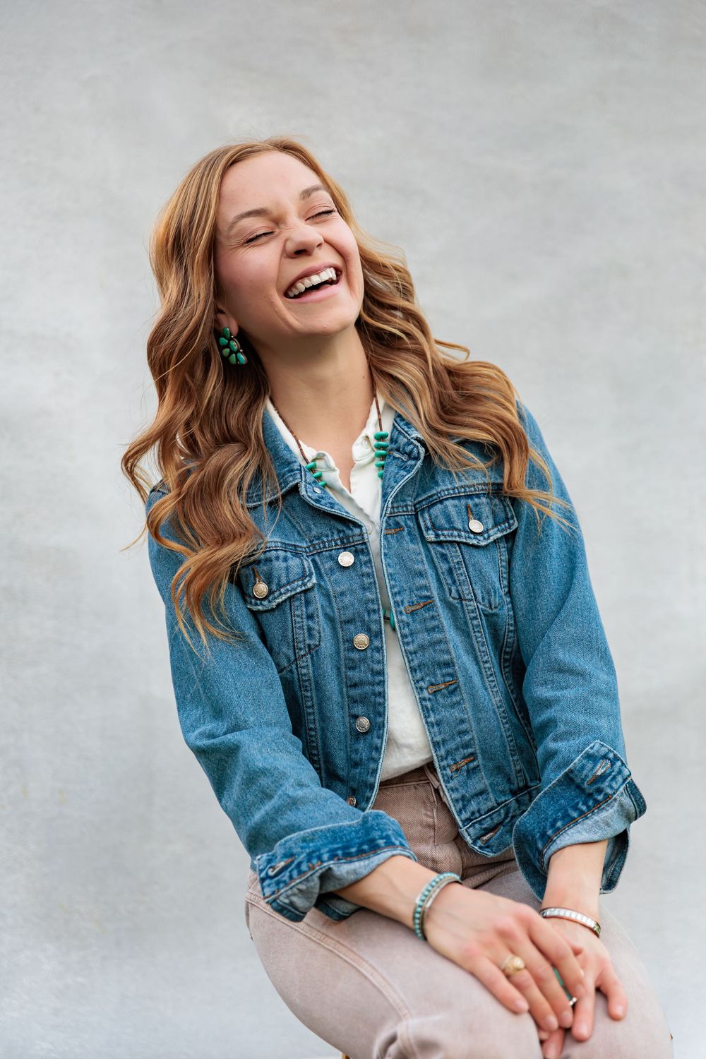 Laughing young woman in denim jacket and green earrings with wavy hair poses against gray wall.