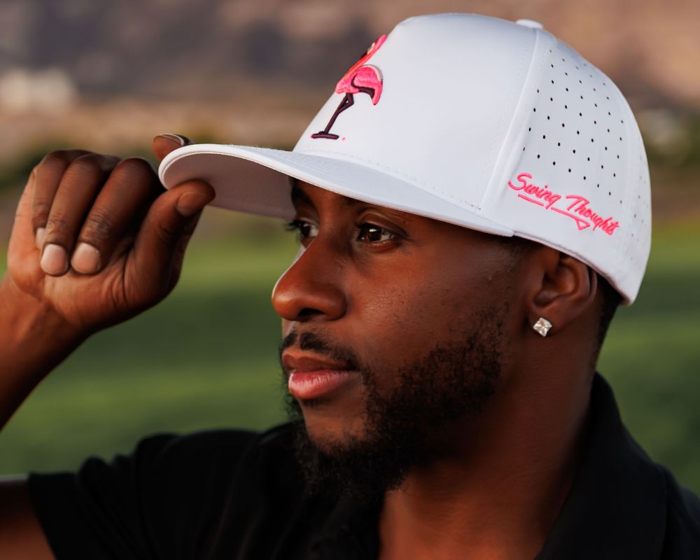 Man wearing a white golf cap with pink flamingo logo and pink Swing Thoughts text, adjusting brim outdoors.