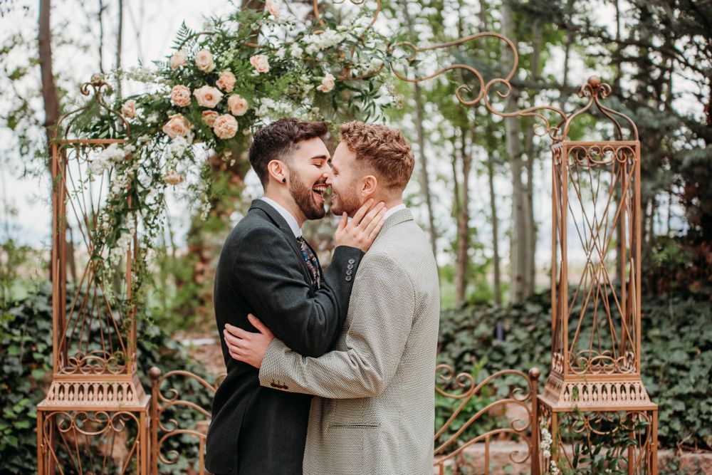 A couple embraces under a floral arch with lanterns in a wooded setting.