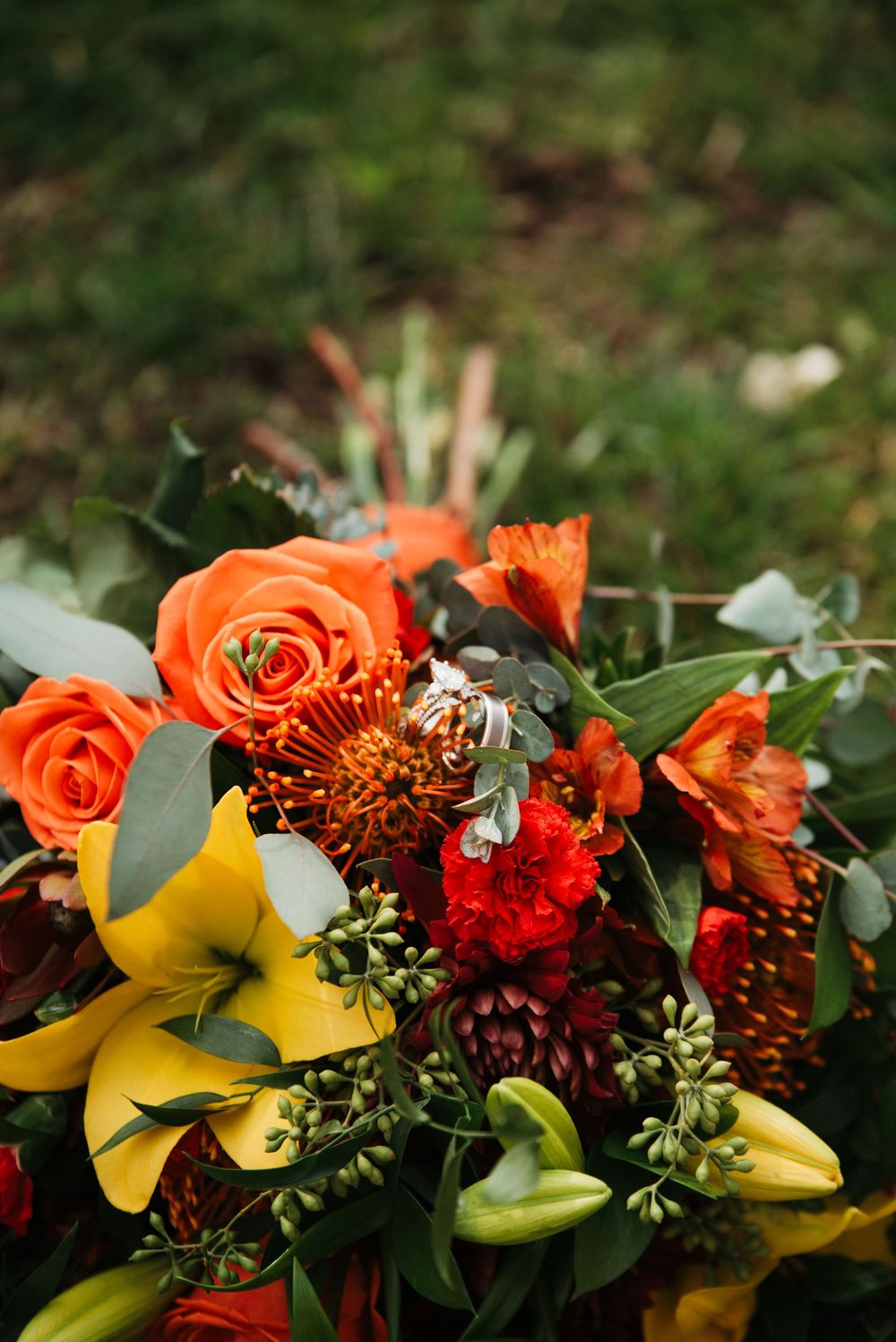 Vibrant fall floral arrangement with orange roses, yellow lilies, and red blooms against green background.
