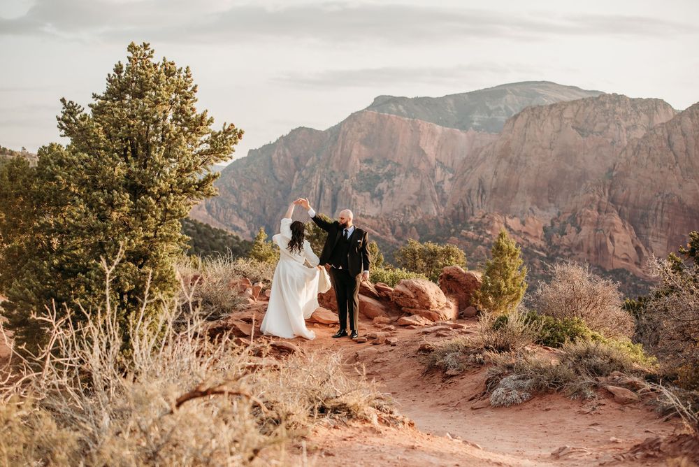 Two people in wedding attire stand on a scenic mountain trail overlooking rugged canyon peaks at sunset.