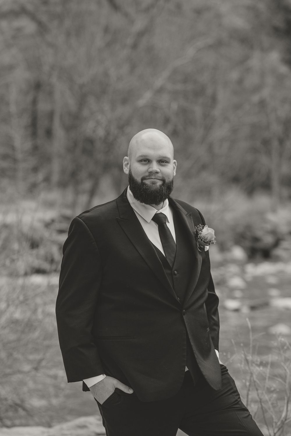 A black and white portrait of a person in a formal suit with a beard standing outdoors.