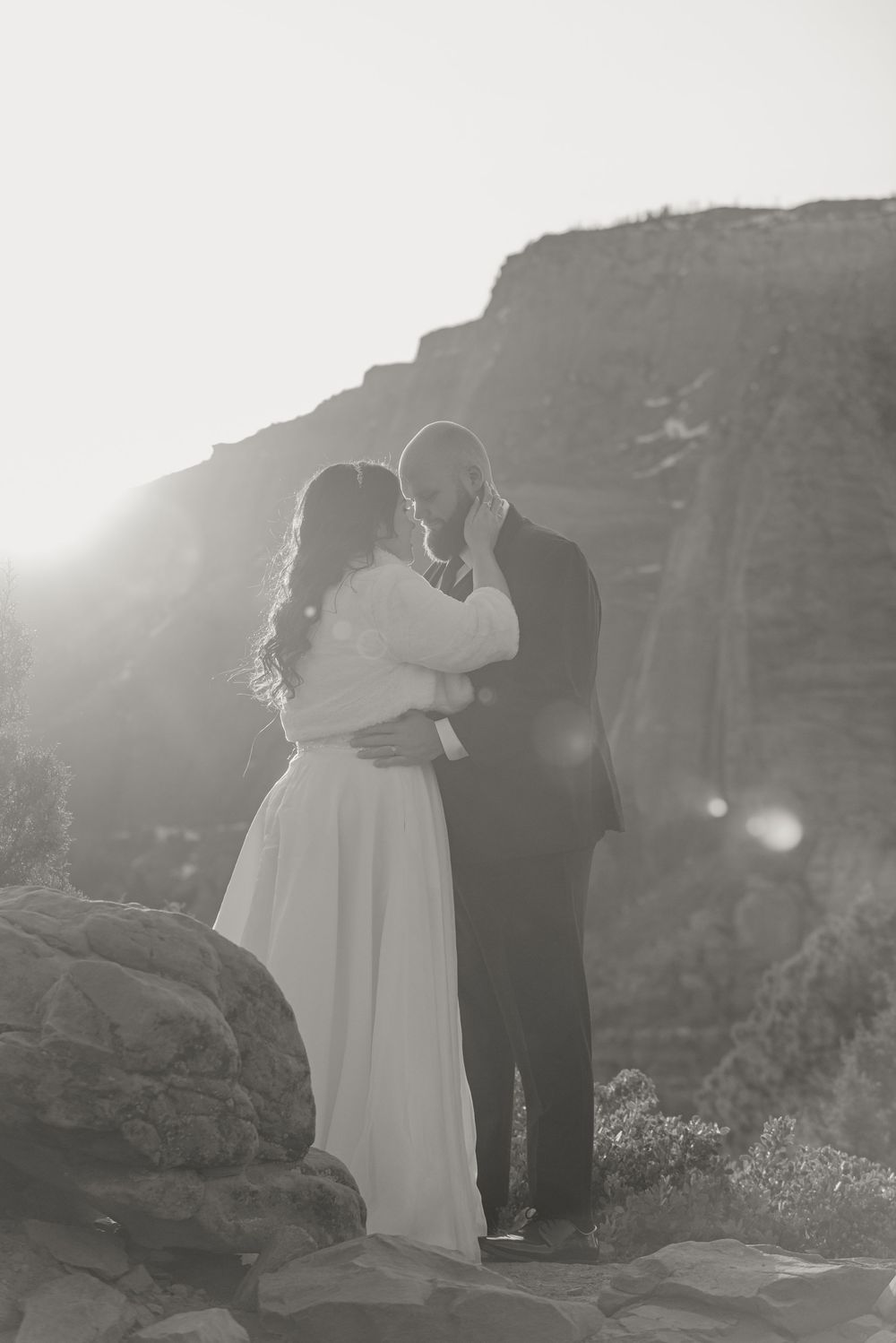 A romantic wedding couple embraces on a rocky cliff with dramatic mountains and sunlight in the background.